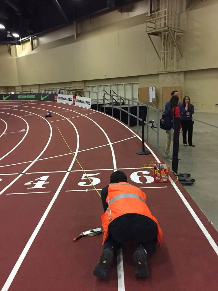 An official in a high-visibility vest checking lane markings on an indoor track.
