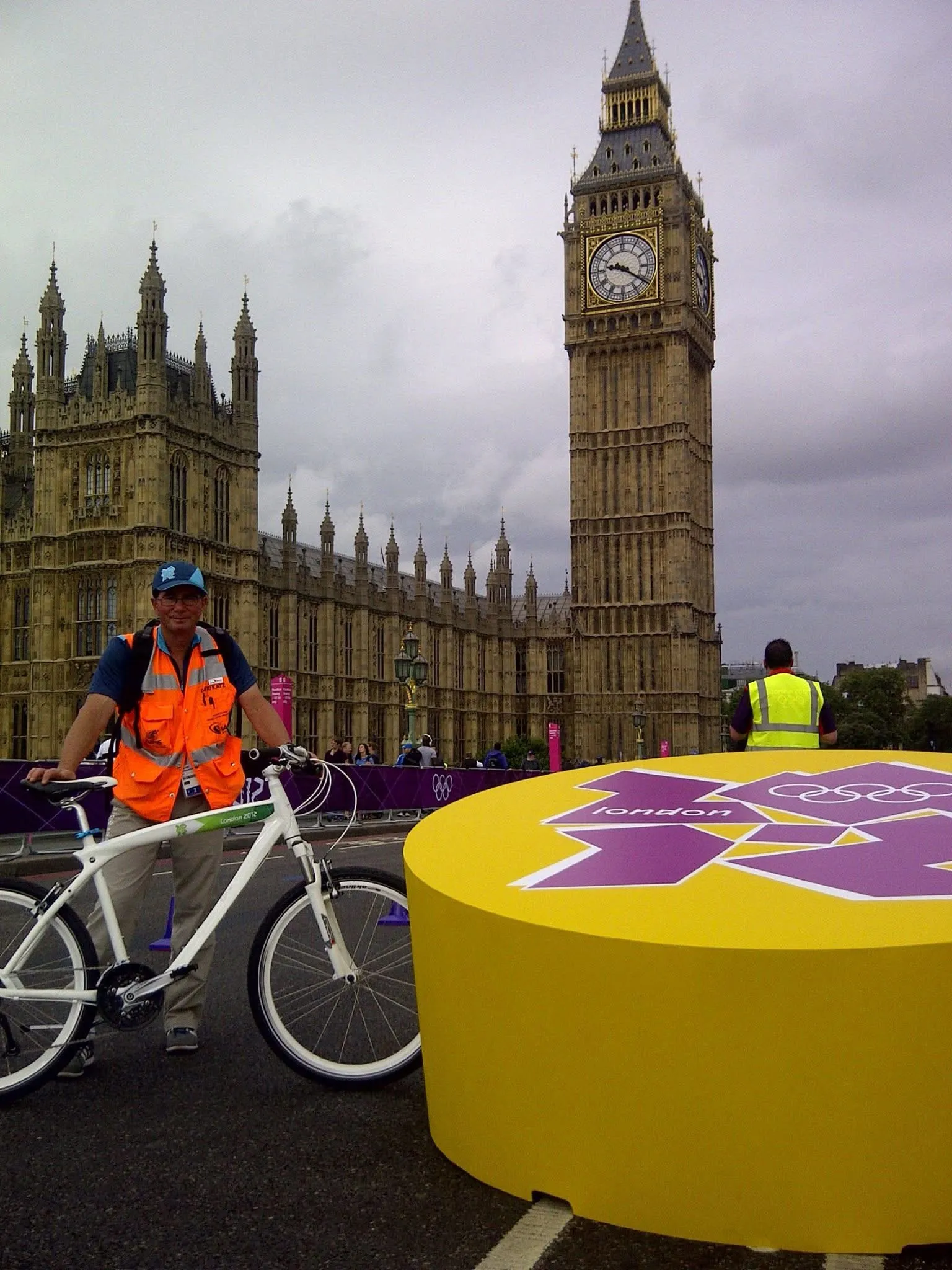 A measurer in an orange vest with a measuring bicycle beside a London 2012 marker, Big Ben behind.