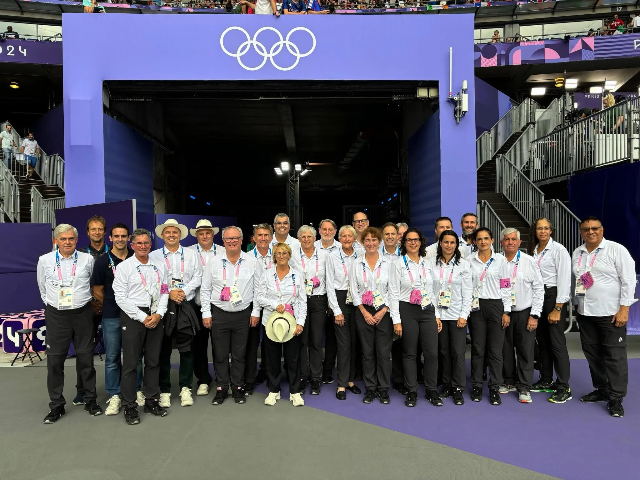 A large group of technical officials in white shirts in front of the Olympic rings.