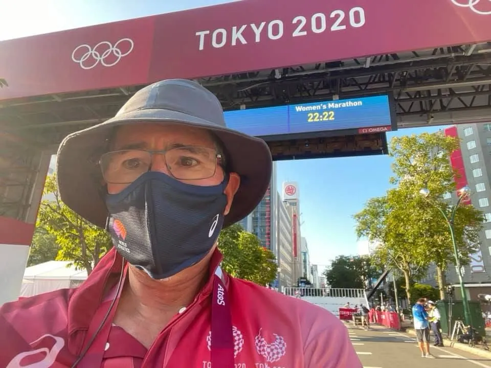 A course official in a mask and sun hat under the Tokyo 2020 marathon gantry.