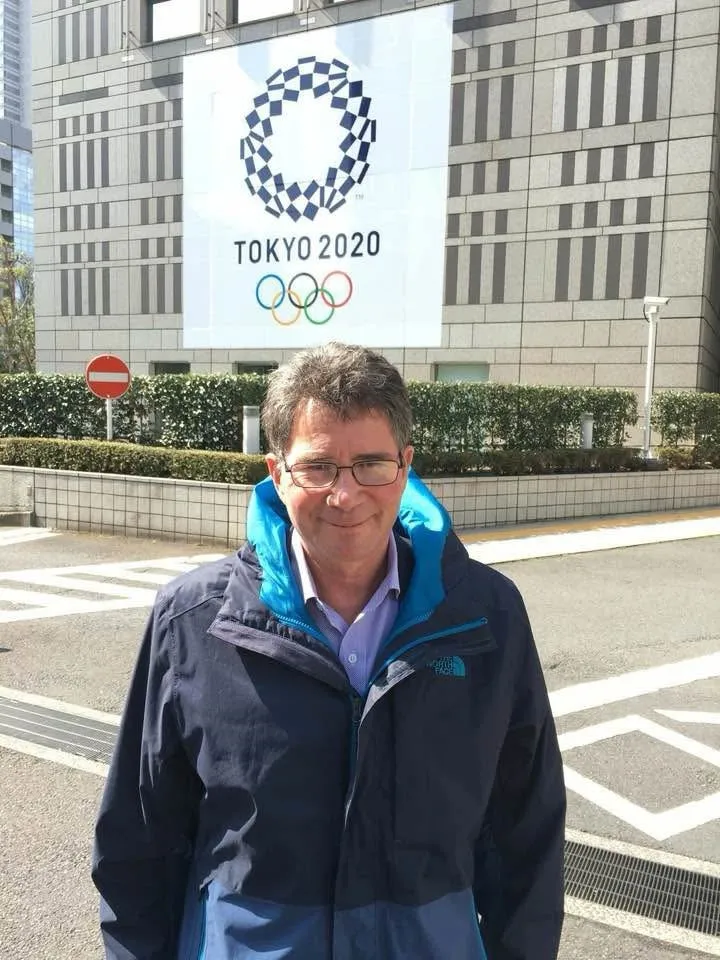 A man in a rain jacket in front of a Tokyo 2020 banner.